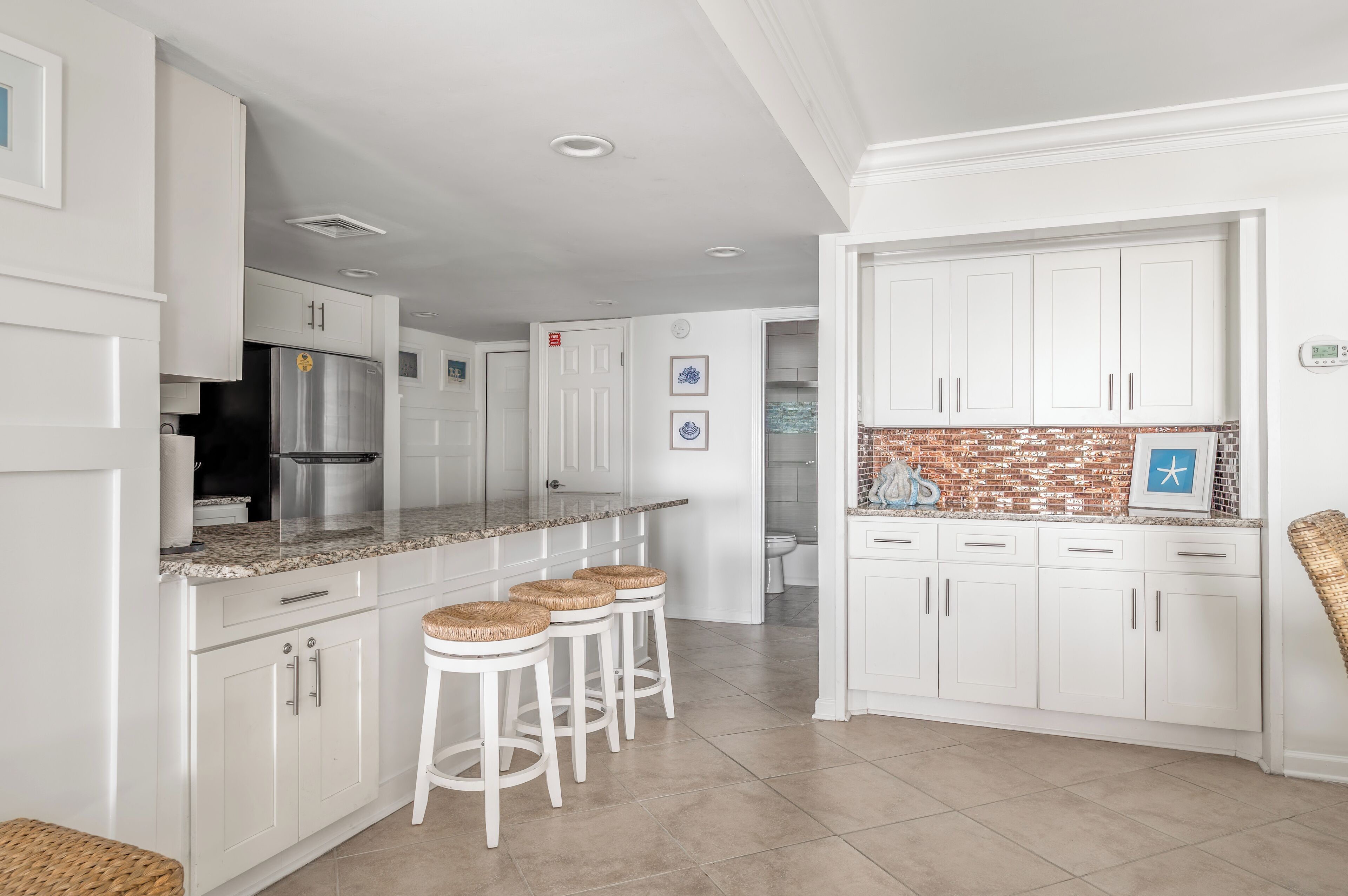 A modern kitchen with white cabinetry, a stainless steel refrigerator, a granite countertop, and three bar stools.