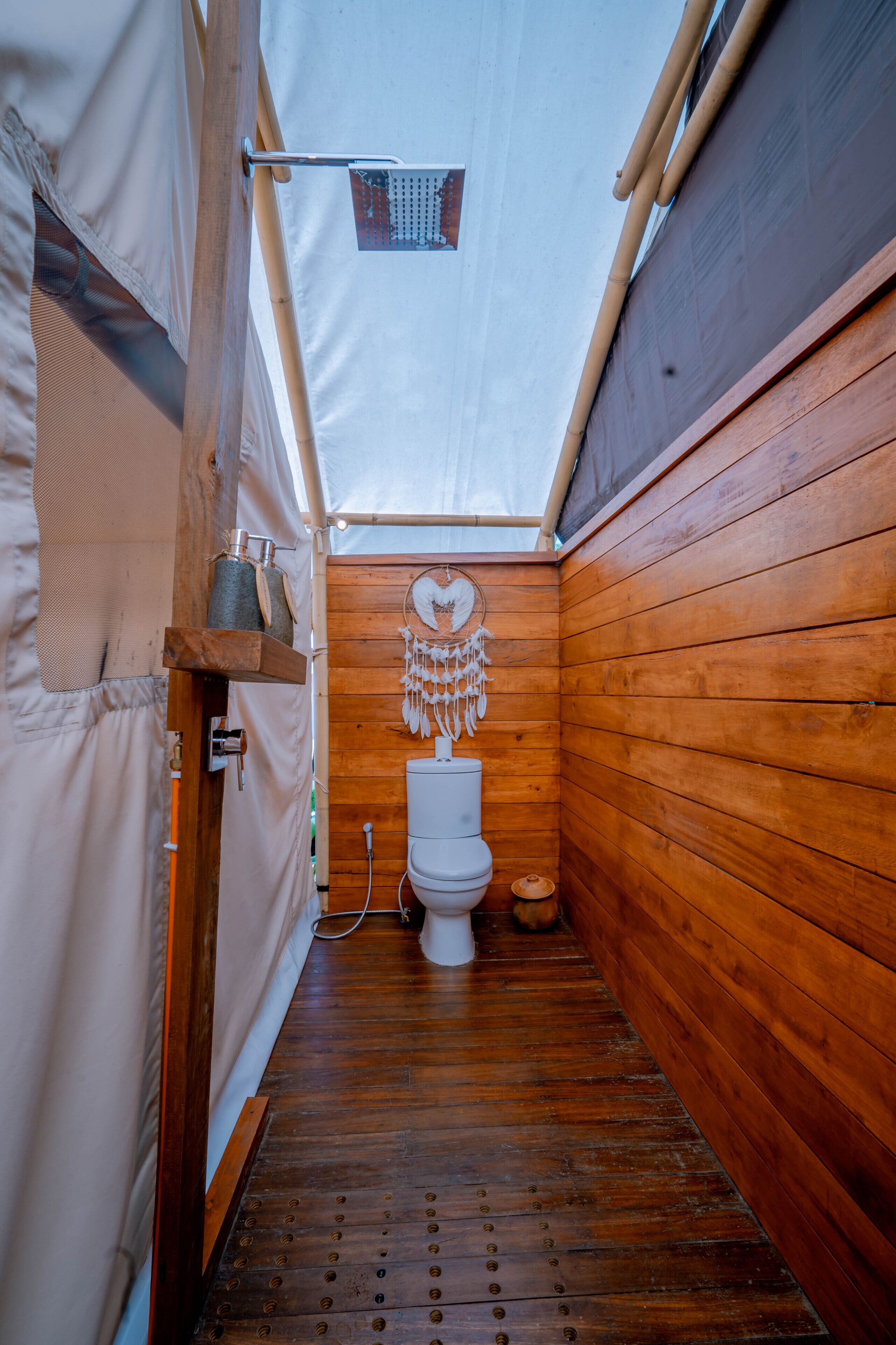 A narrow wooden bathroom with a white toilet, a hanging dreamcatcher, and a small shelf with a container.