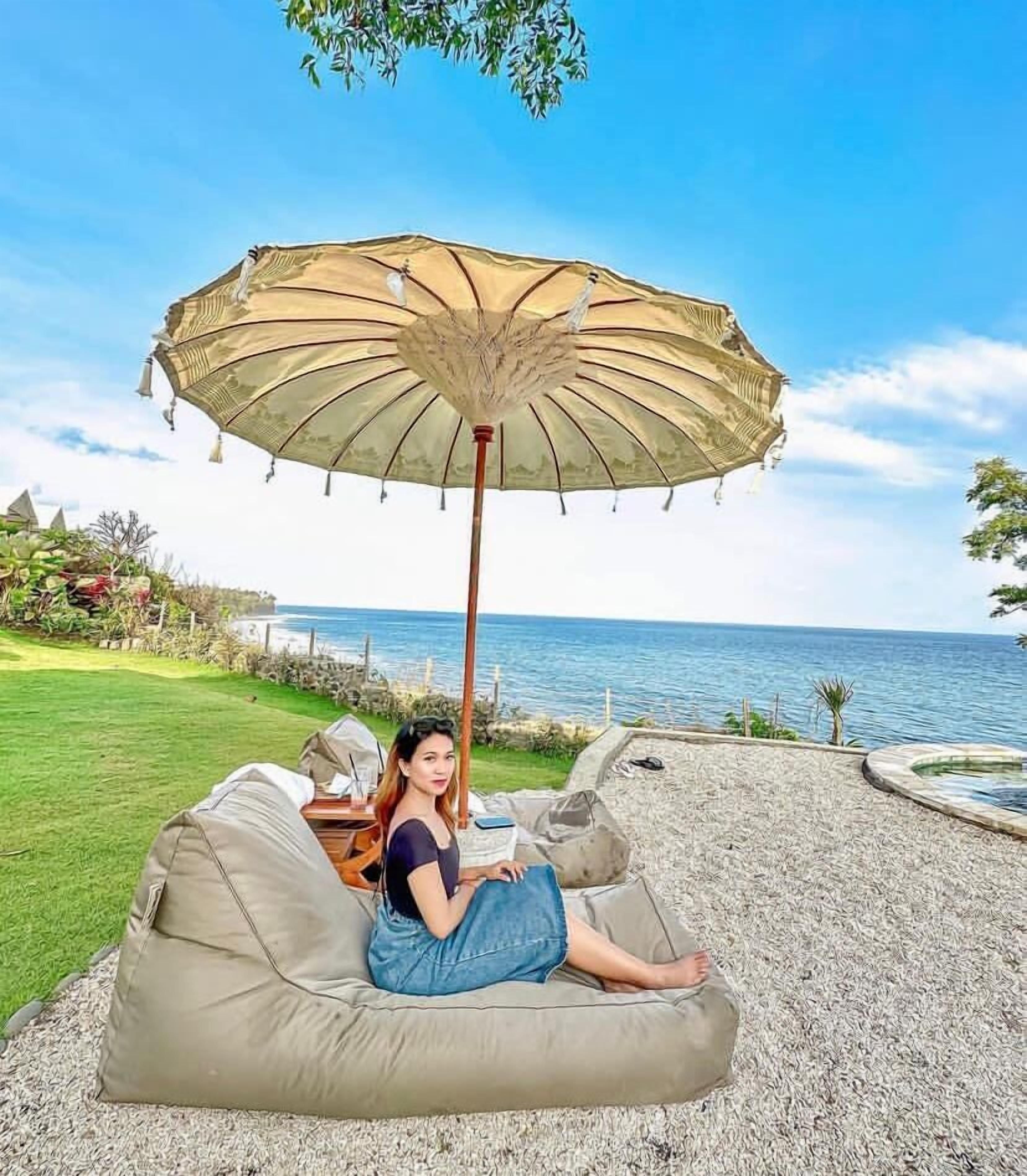 A person sitting on a beige outdoor sofa with a large umbrella, overlooking a body of water.