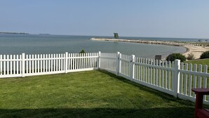 A view of a beach with a white picket fence, a grassy lawn, and a rocky breakwater in the distance.