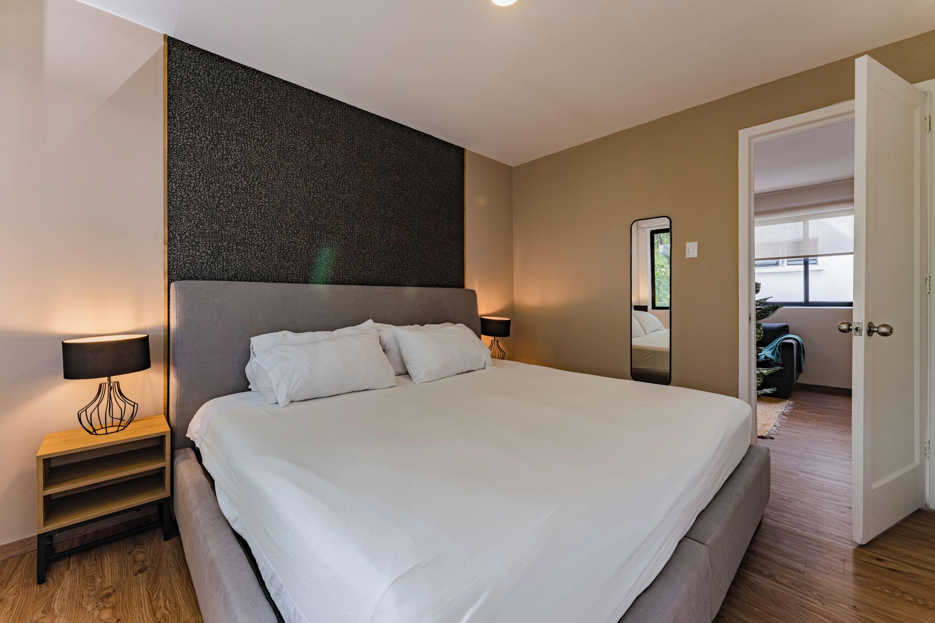 A neatly made bed with white linens, a small bedside table with a lamp, and a large mirror on the wall.