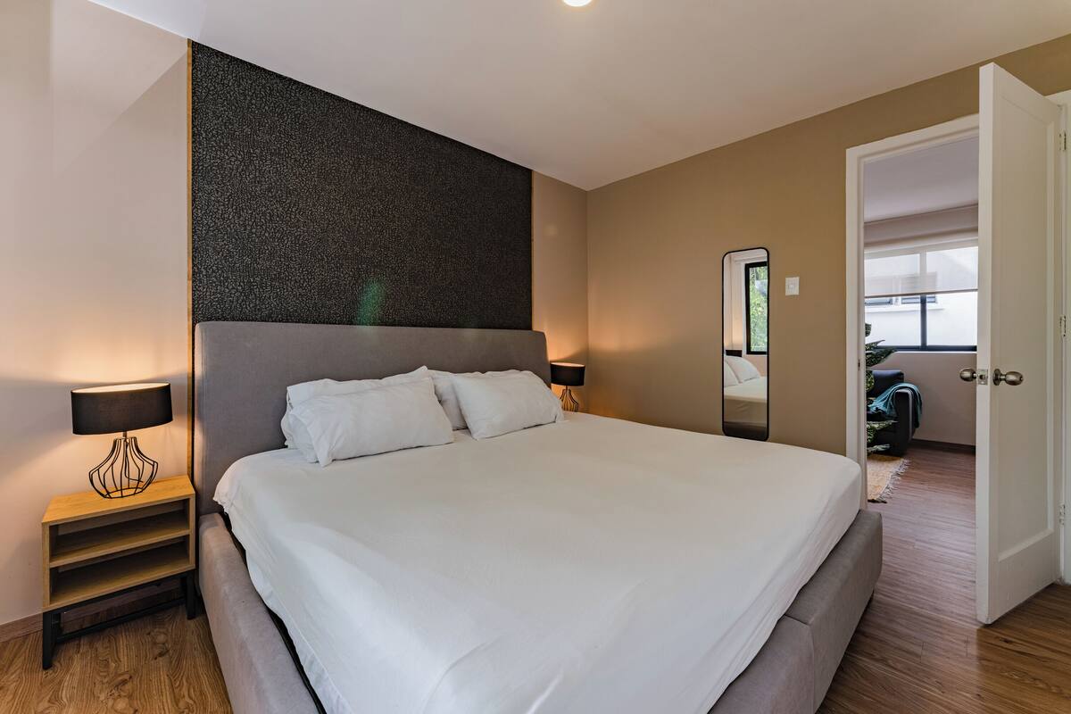 A neatly made bed with white linens, a small bedside table with a lamp, and a large mirror on the wall.