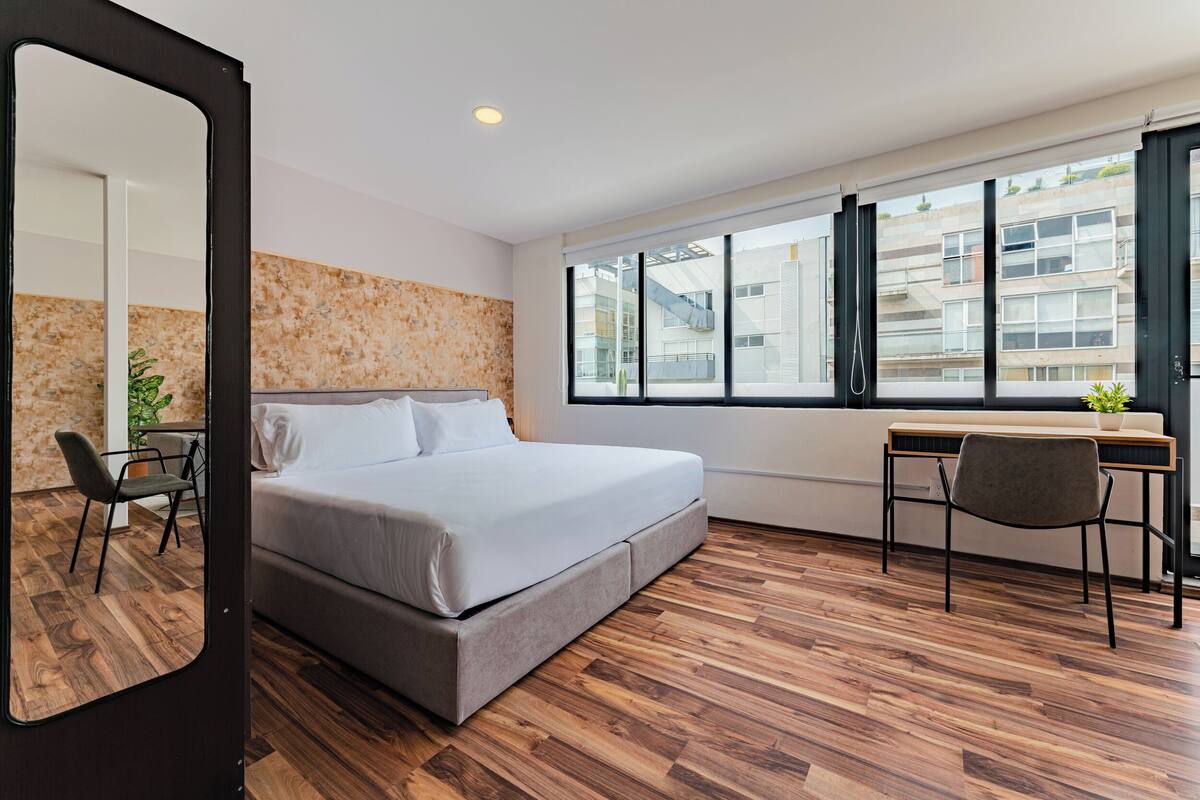 A modern bedroom with a large bed, a desk, and a chair. There is a window with a view of buildings outside.