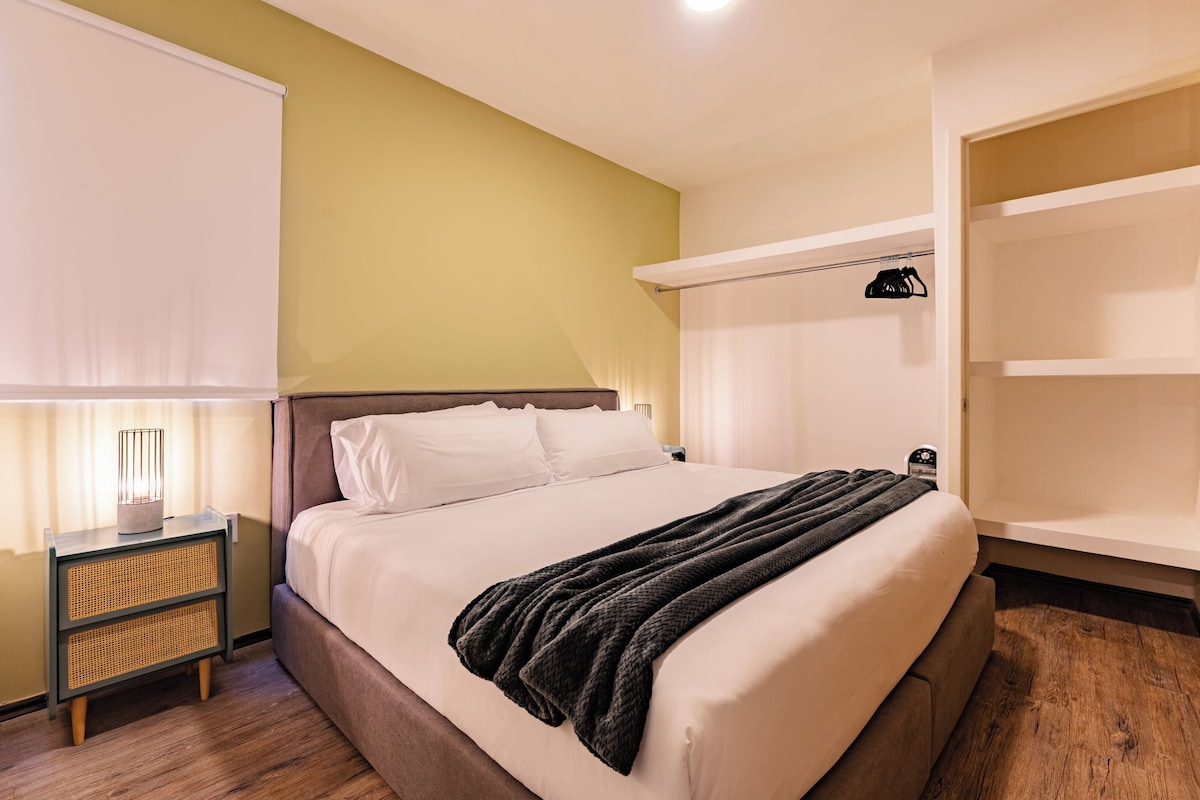 A neatly made bed with a white comforter and a dark blanket, a bedside lamp, a nightstand with a woven side table, and a built-in wardrobe with a hanging rod.
