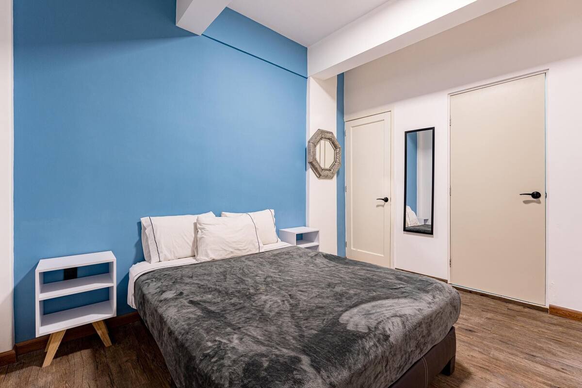 A bedroom with a bed, white nightstands, a blue accent wall, a door, and a mirror.