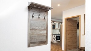 A hallway with a rustic wooden wall-mounted rack with hooks, a wooden door, and a kitchen area in the background.