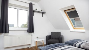 A modern bedroom with a sloped ceiling, a large window with curtains, a radiator, a small table with a plant, a grey armchair, and a bed with striped bedding.