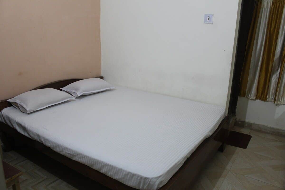Uma cama de solteiro com colchão branco e duas almofadas, cabeceira de madeira e parede lisa com tomada.