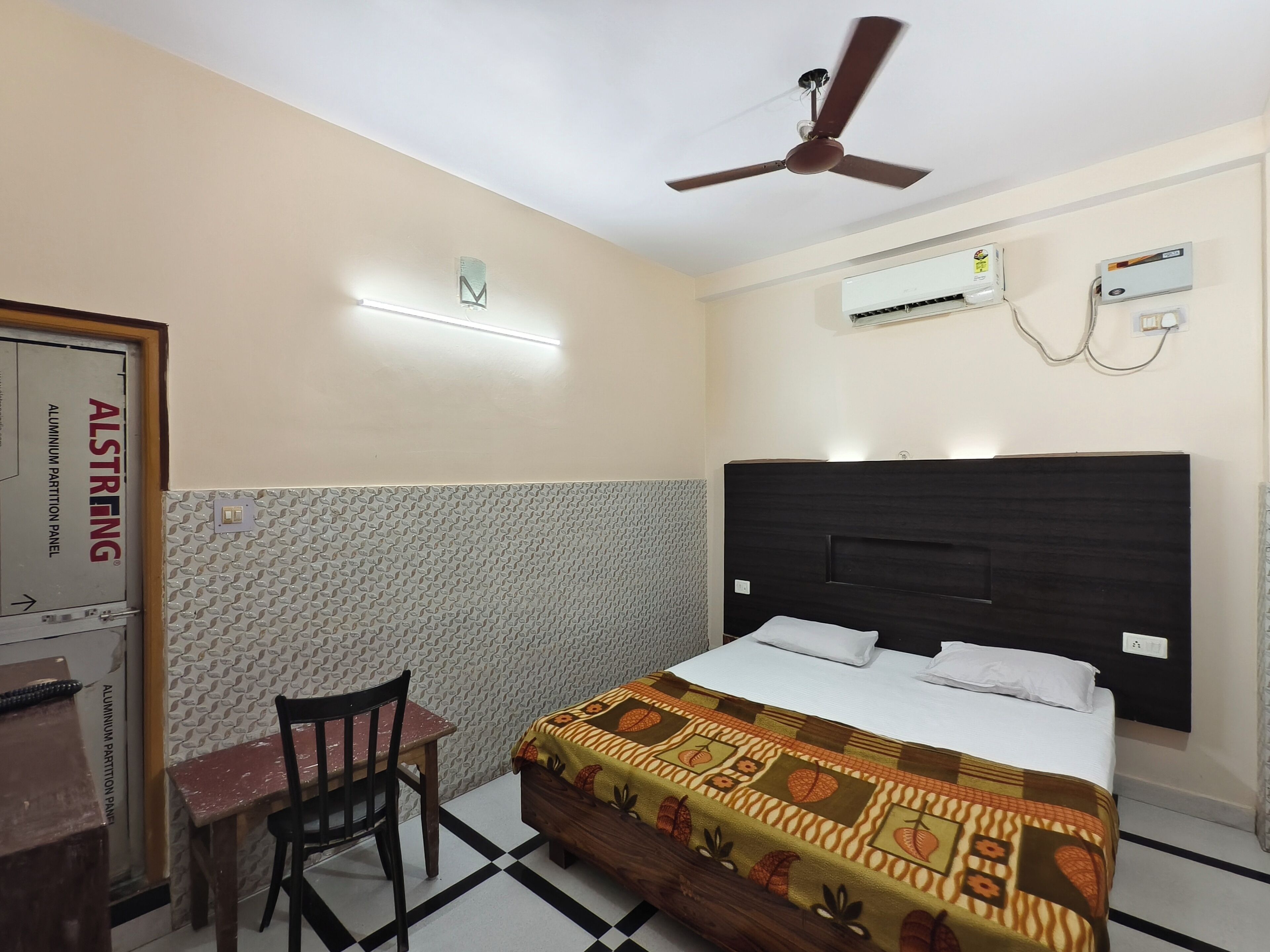 Une chambre d’hôtel comprenant un lit, un bureau, une chaise, un ventilateur de plafond, un appareil de climatisation et un mur à texture.