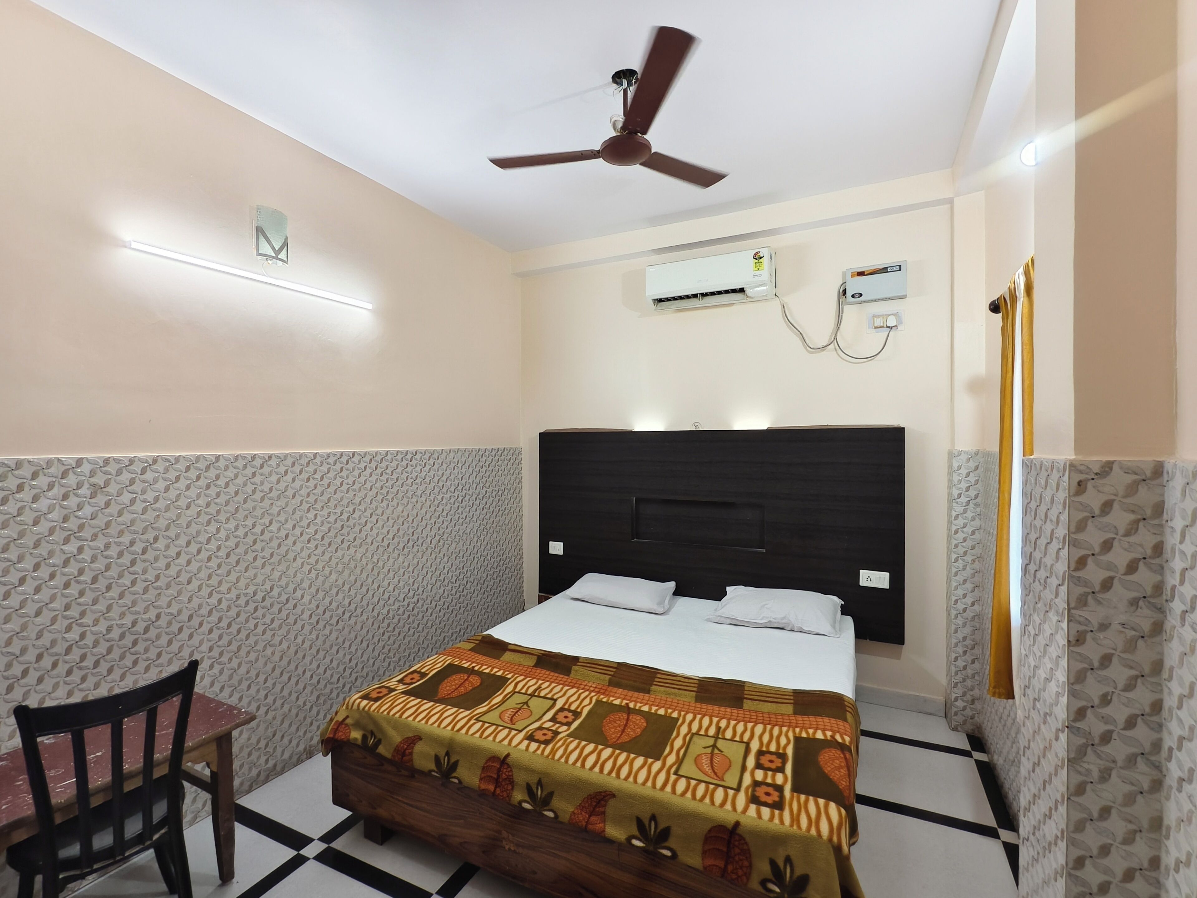 A hotel room with a bed, a desk, a chair, a ceiling fan, and two wall-mounted air conditioners.