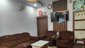 A living room with a brown sofa set, a white coffee table, a flat-screen TV mounted on a wooden panel, and a patterned wall hanging. - Hotel Vijay (Rajgir)