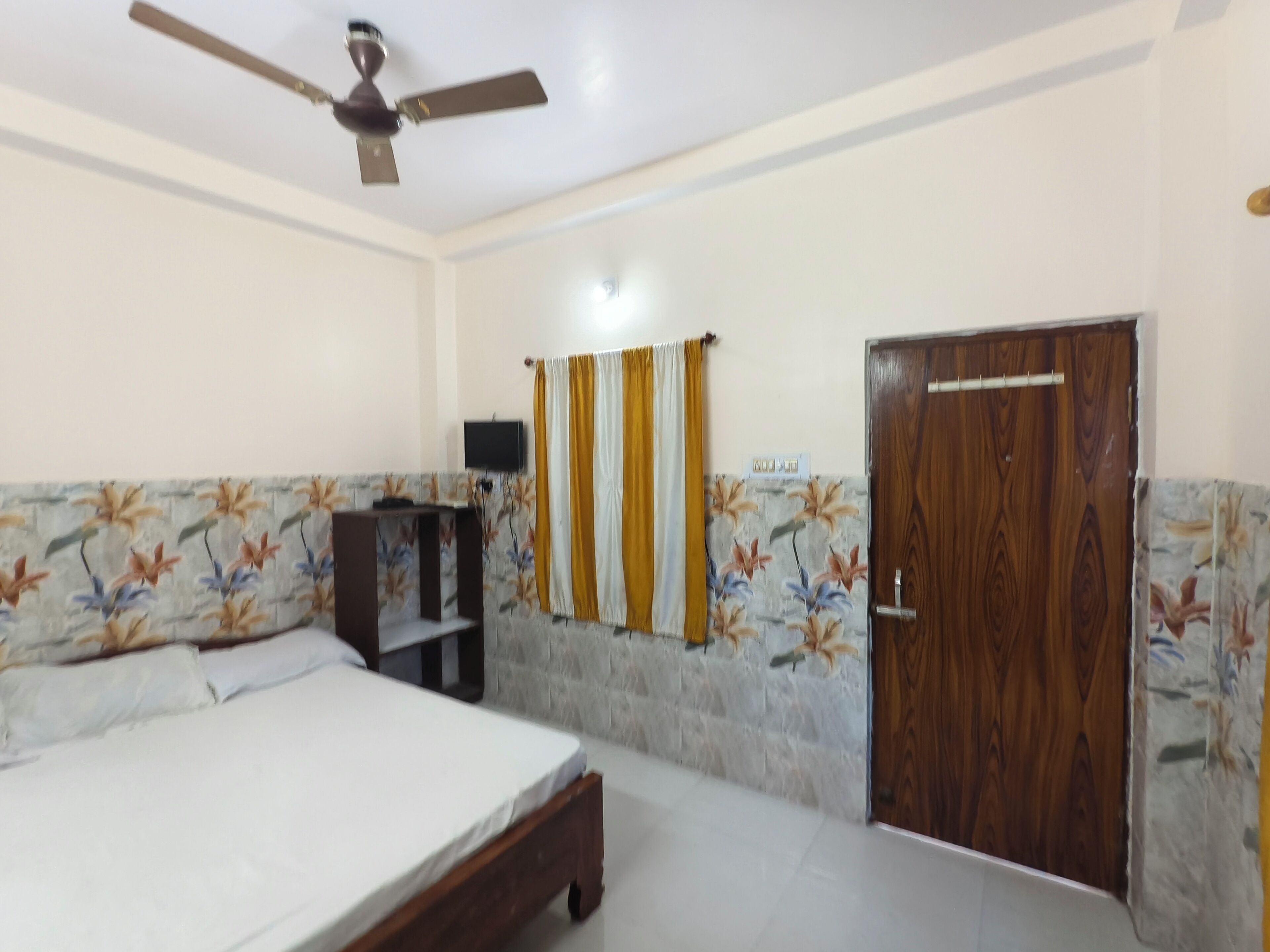 A bedroom with a bed, a ceiling fan, a wooden door, and a wall-mounted TV.