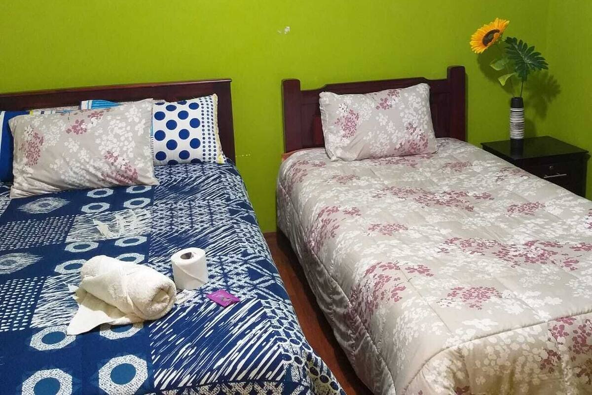 A room with two beds, one with a blue patterned bedspread and the other with a floral bedspread. There is a framed picture on the wall above the beds, a small table with a vase of flowers, and a towel on the floor.
