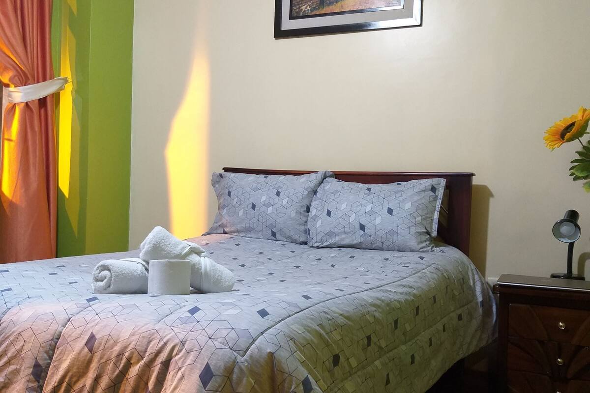 A hotel room with a bed, a nightstand, a lamp, a framed painting, and a window with curtains.
