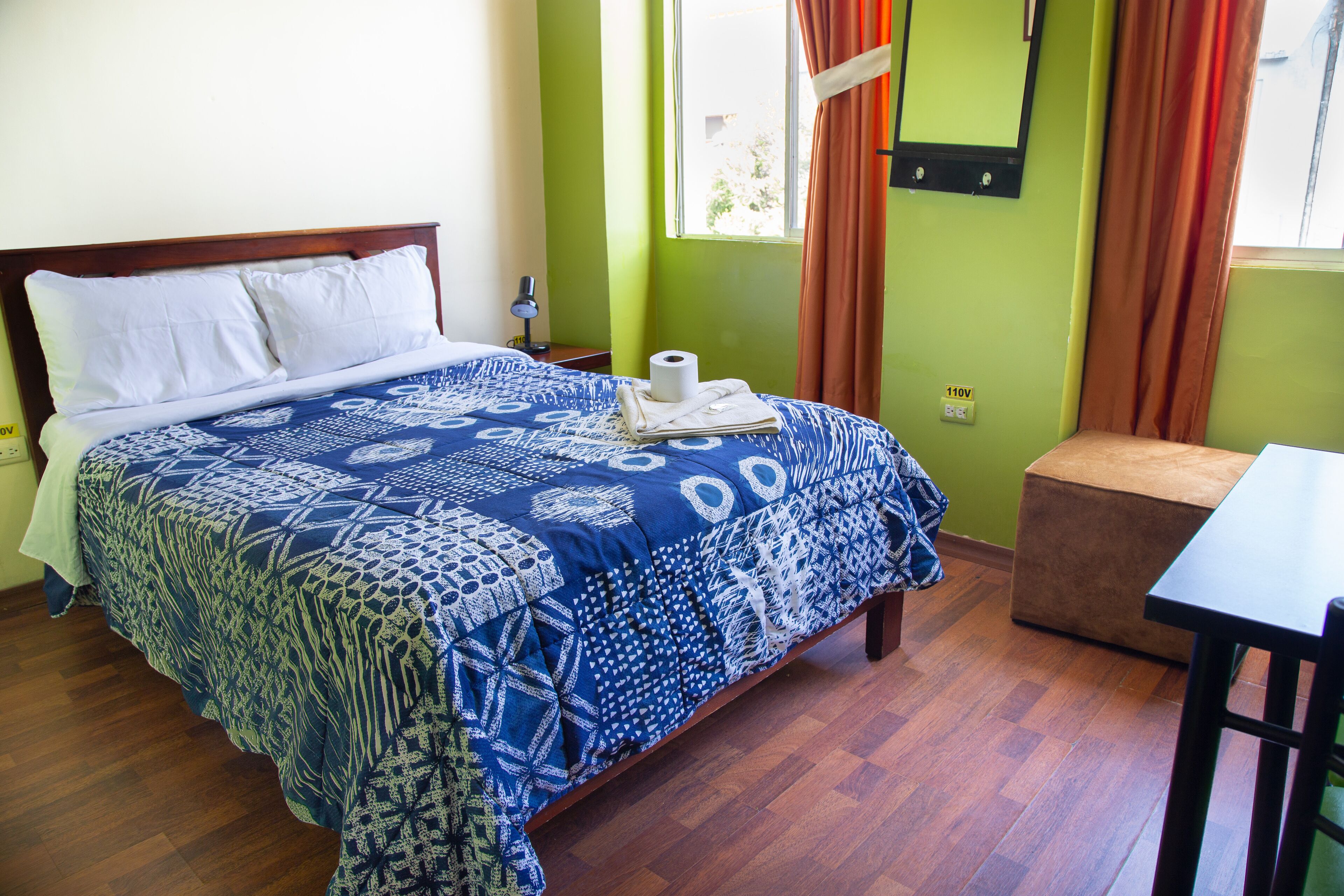 A neatly made bed with a patterned bedspread, a bedside table with a lamp, and a window with curtains.