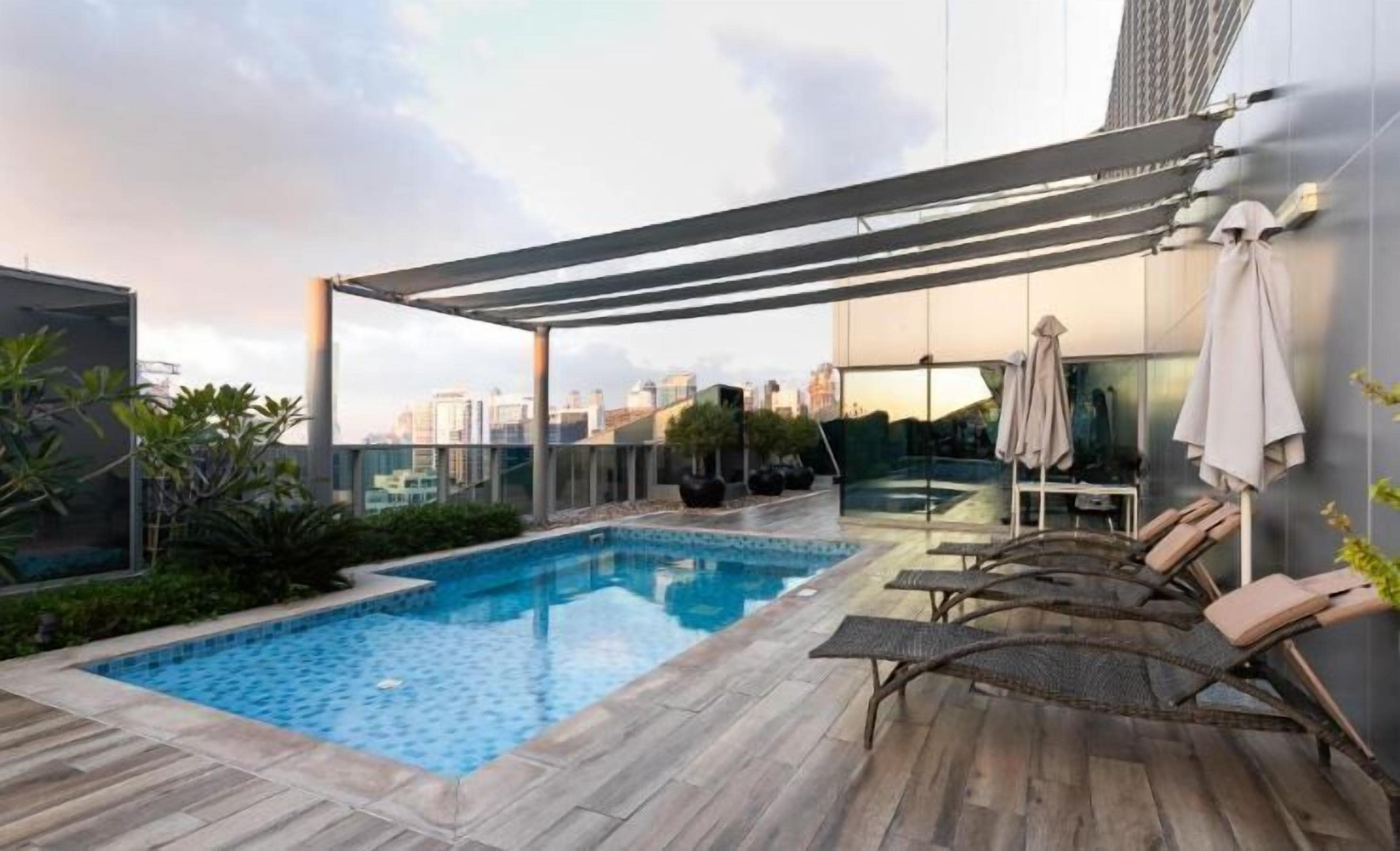 A rooftop pool area with a clear view of the city skyline, lounge chairs, and a pergola providing shade.