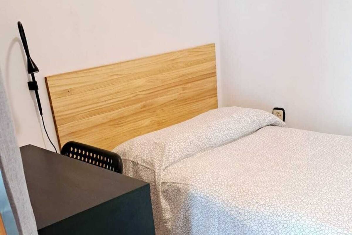 A small, neatly arranged bedroom with a wooden headboard, a single bed with a patterned bedspread, and a black desk with a chair.