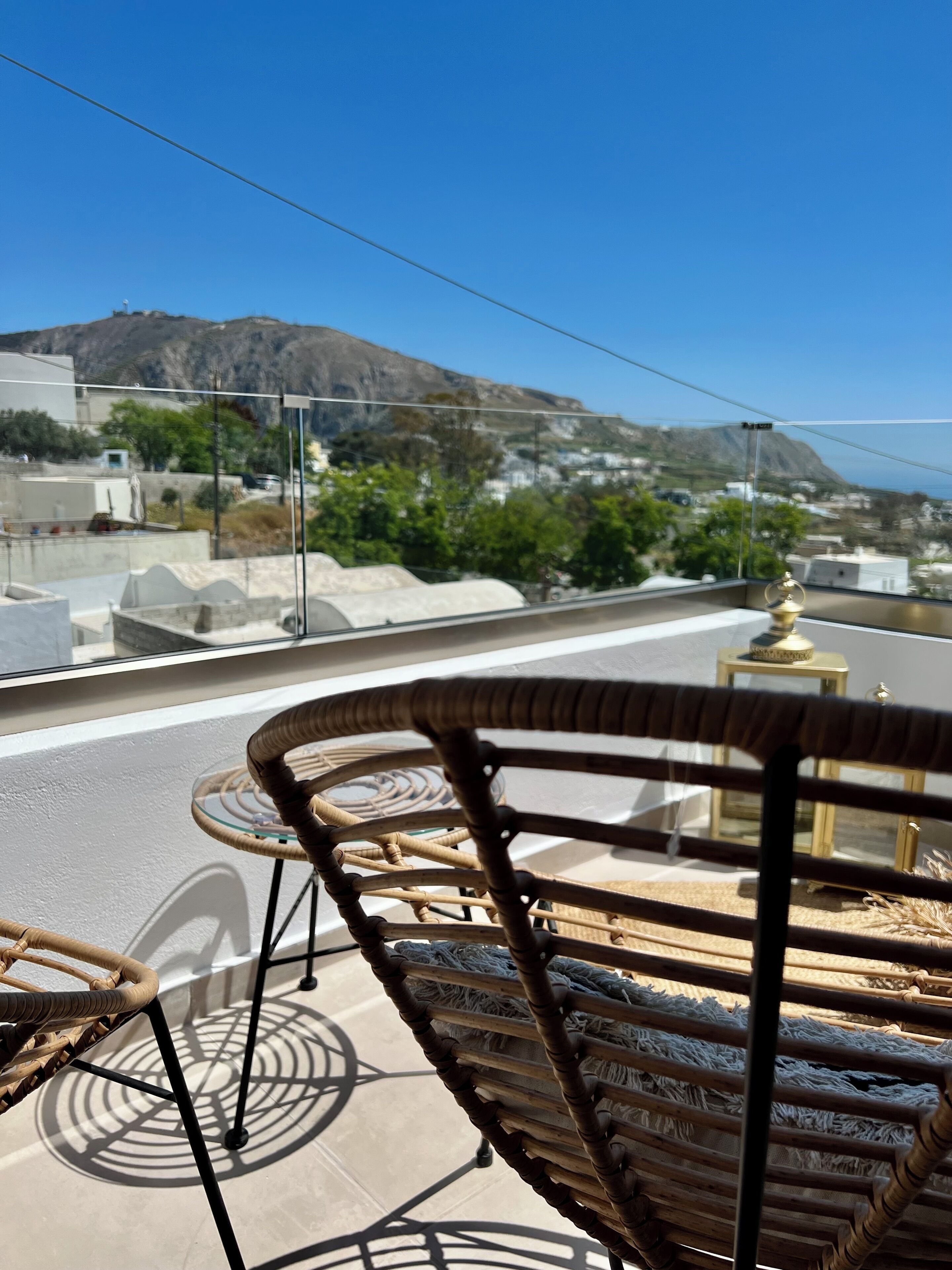 A balcony with wicker chairs and a small table, overlooking a coastal town with buildings and a mountain in the background.