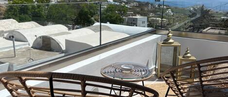 A terrace with wicker chairs and a glass table, overlooking a coastal landscape with buildings and hills.