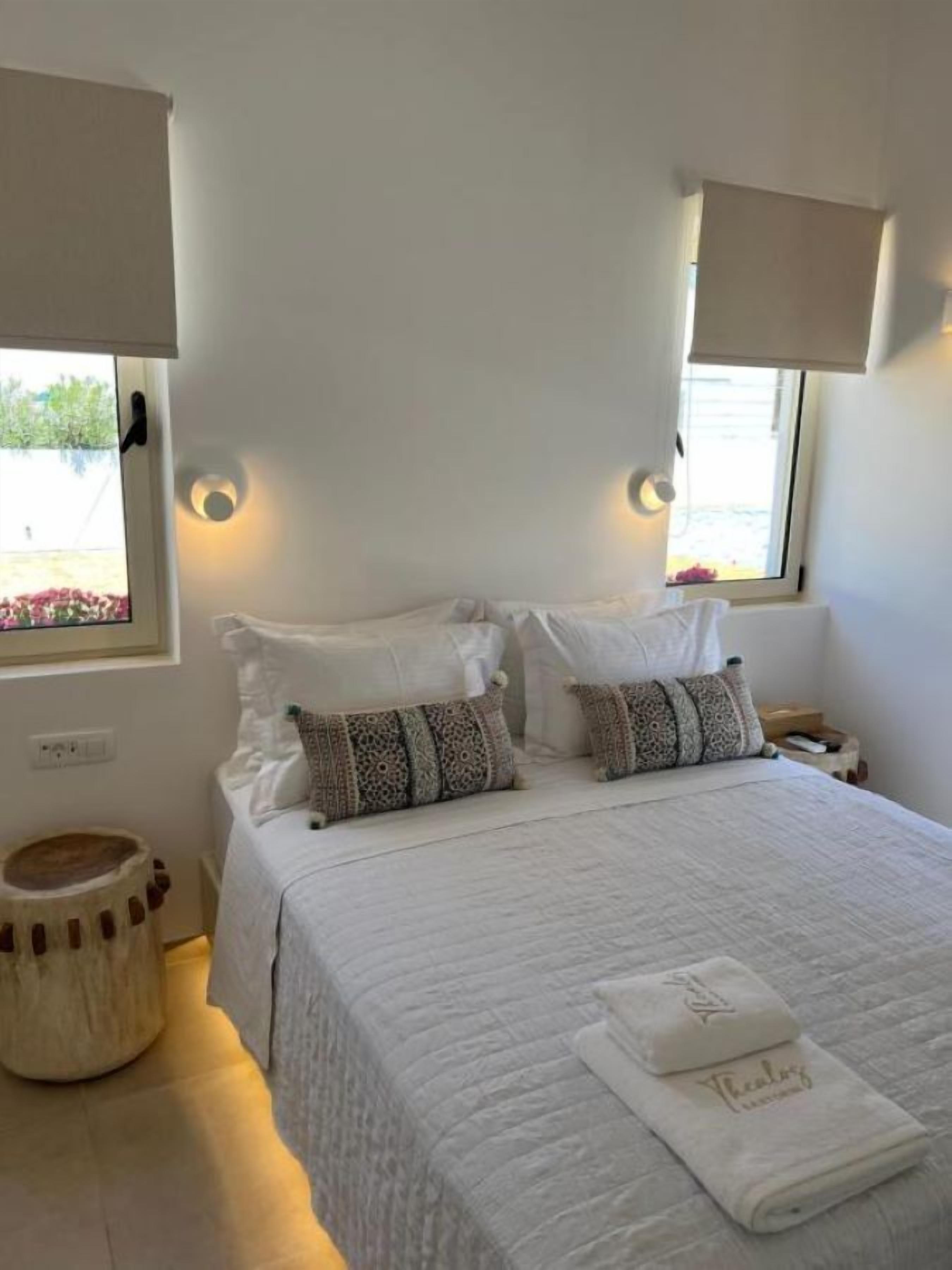 A double bed with white bedding and patterned pillows, two bedside tables with lamps, a small round stool, and a window with a view of greenery.