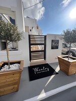 Un edificio moderno con un portón negro, macetas de madera con guijarros blancos y un felpudo negro con la inscripción "Thecalos Santorini".