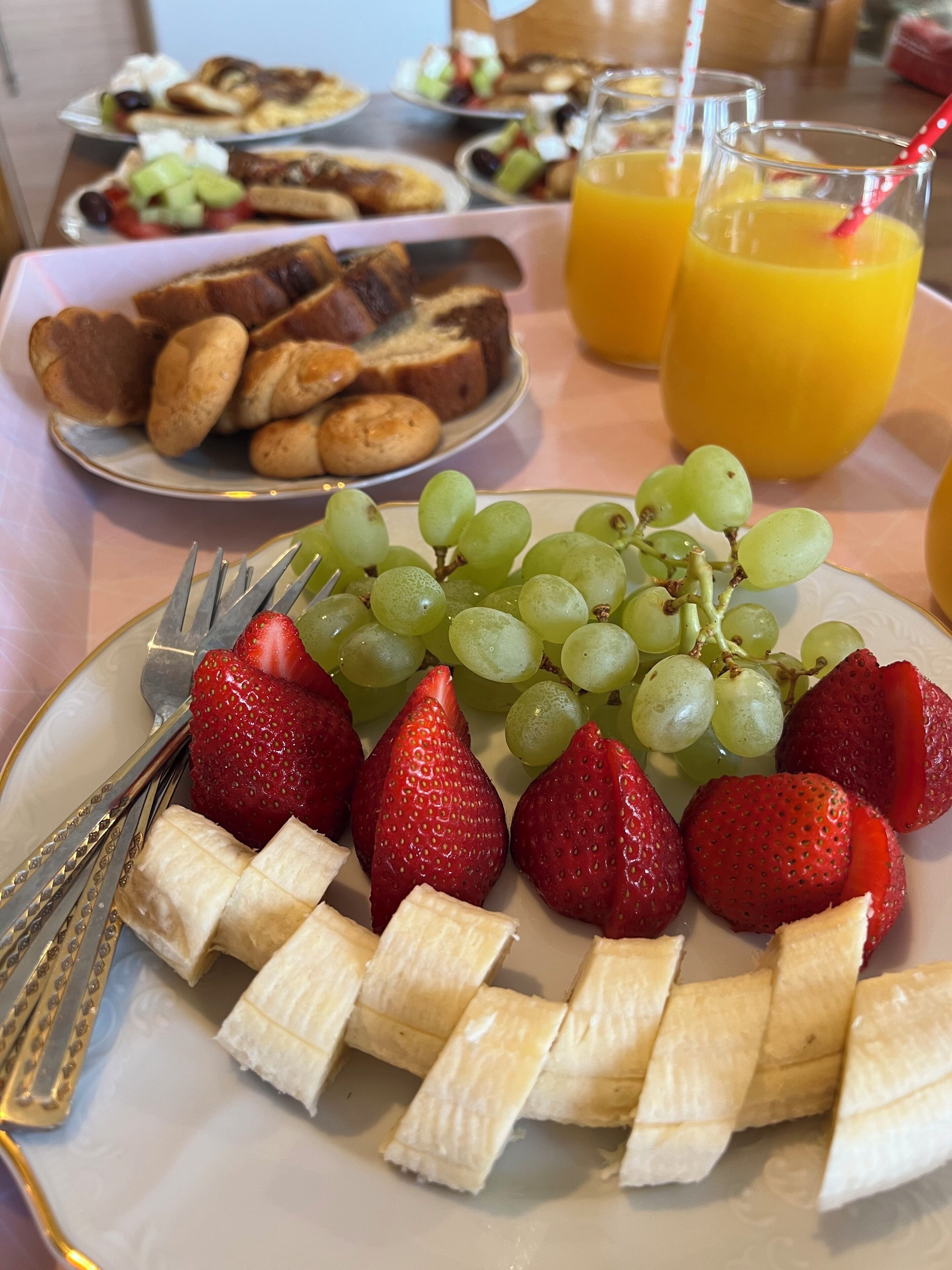Desayuno con un plato de rodajas de plátano, fresas y uvas verdes, acompañado de tostadas y vasos de jugo.