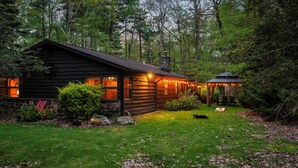 A log cabin with a stone chimney, surrounded by a well-maintained lawn and dense forest. - Ranch on Greenwood: Hot tub , fire pits, kayaks, Rafts, Bikes (Lake Harmony)