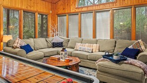 A cozy living room with a sectional sofa, wooden walls, large windows, and a coffee table with board games. - Ranch on Greenwood: Hot tub , fire pits, kayaks, Rafts, Bikes (Lake Harmony)