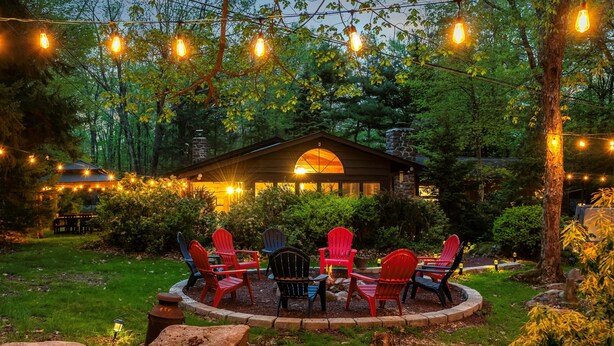 A cozy outdoor seating area with red and blue Adirondack chairs, surrounded by a stone fire pit, under string lights. - Ranch on Greenwood: Hot tub , fire pits, kayaks, Rafts, Bikes (Lake Harmony)