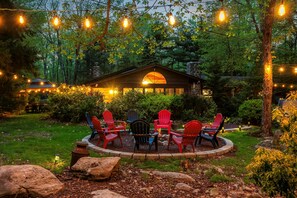 A cozy outdoor seating area with red and blue Adirondack chairs, surrounded by a stone fire pit, under string lights. - Ranch on Greenwood: Hot tub , fire pits, kayaks, Rafts, Bikes (Lake Harmony)