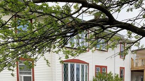 Une maison blanche aux bordures rouges, avec un porche abrité et trois chaises Adirondack bleues disposées sur une pelouse verdoyante.