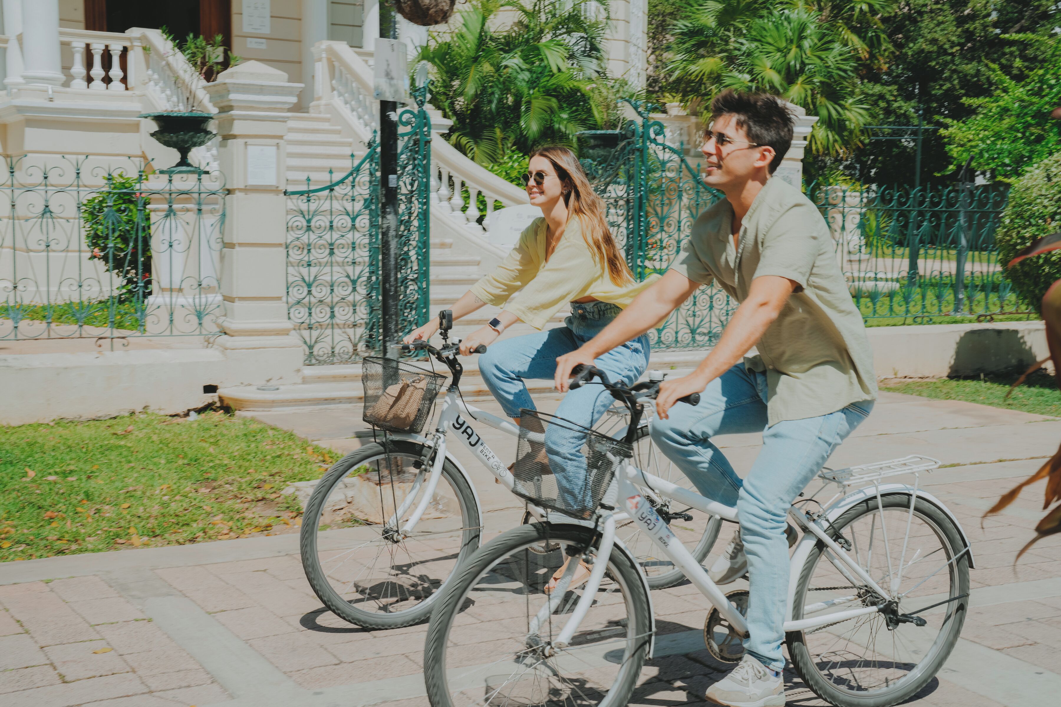 Dos personas montando en bicicleta sobre un camino pavimentado, con un edificio blanco y vallas verdes al fondo.