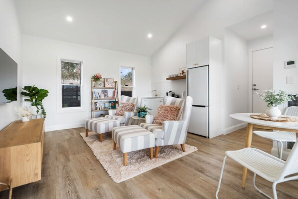 A modern kitchen with white cabinetry, a built-in oven, and a refrigerator. There is a dining area with a white table and chairs, and a cozy seating area with striped chairs and a fluffy rug. - Peaceful & Cosy Studio - Immaculate Presentation (Orange)