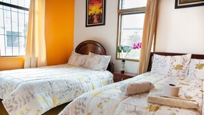 A hotel room with two beds, a window with curtains, a painting on the wall, and a view of the outside. - Hostal Whipala (Quito)