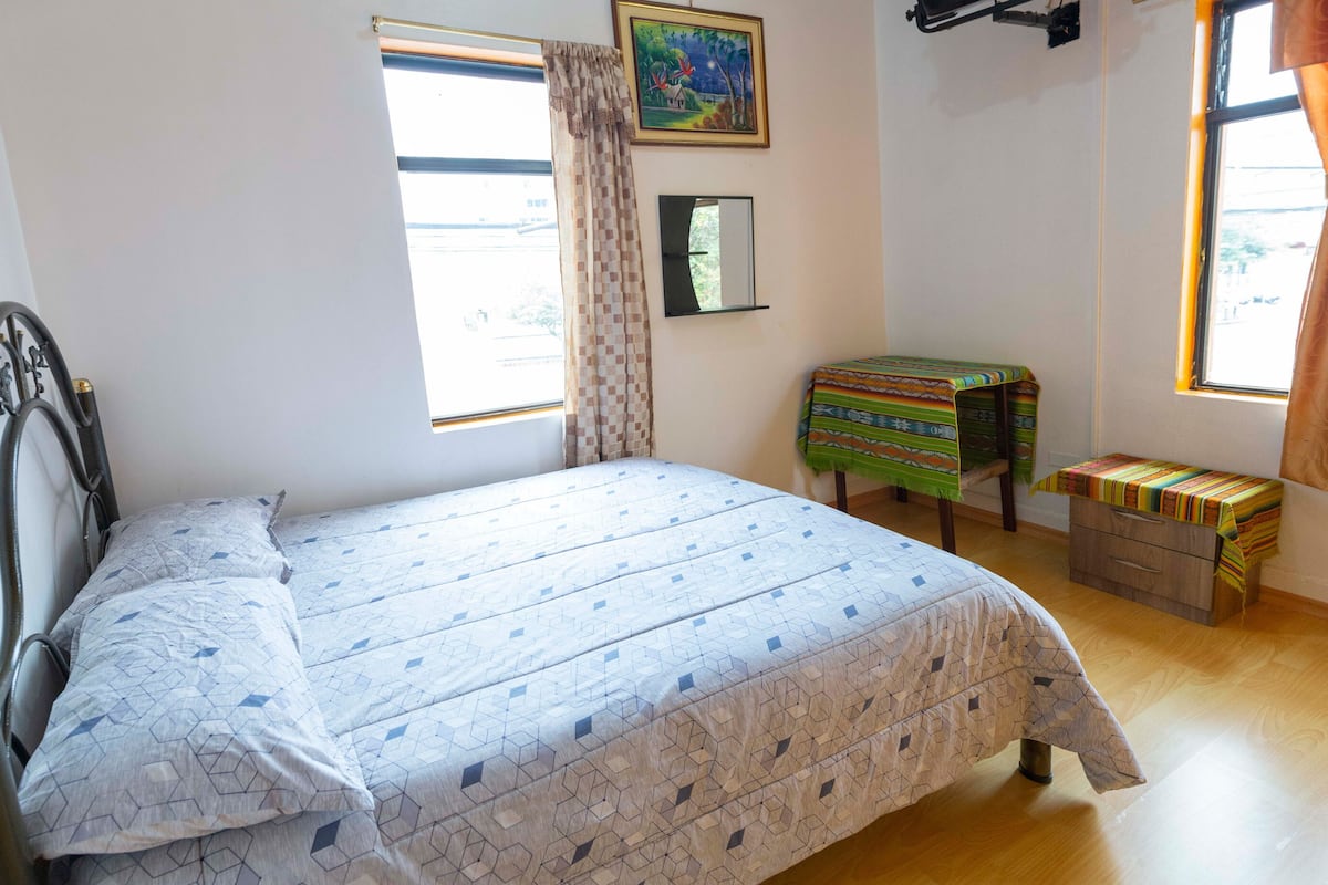 A bedroom with a bed, a small table, a bench, and two windows with curtains.