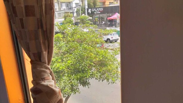 A window with a curtain partially drawn, revealing a street view with buildings and a tree. - Hostal Whipala (Quito)