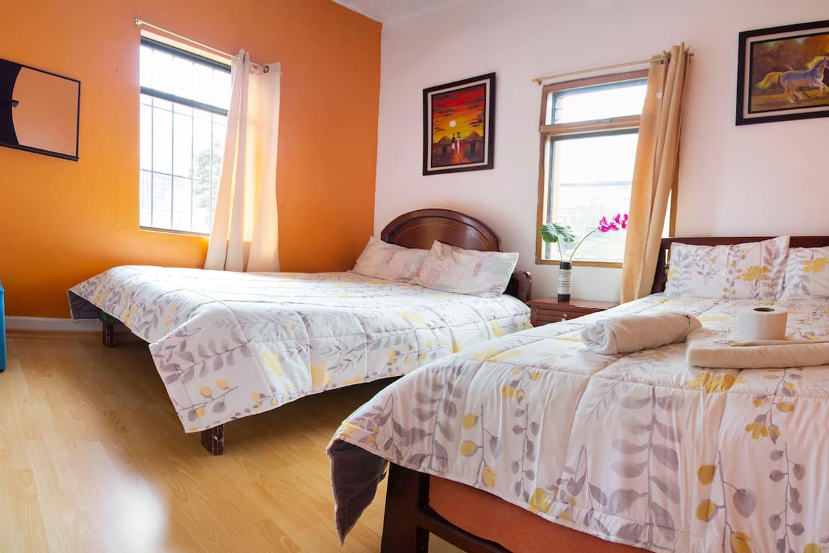 A hotel room with two beds, a blue chair, a wooden floor, and a window with curtains.