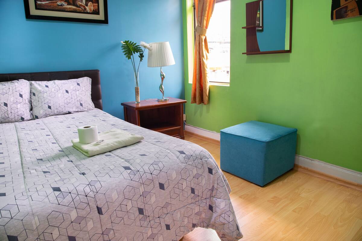 A bedroom with a bed, a nightstand, a lamp, a window with curtains, and a blue ottoman.