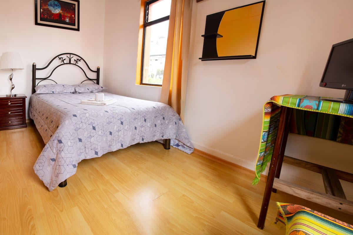 A bedroom with a bed, a nightstand, a lamp, a TV, and a window with curtains.