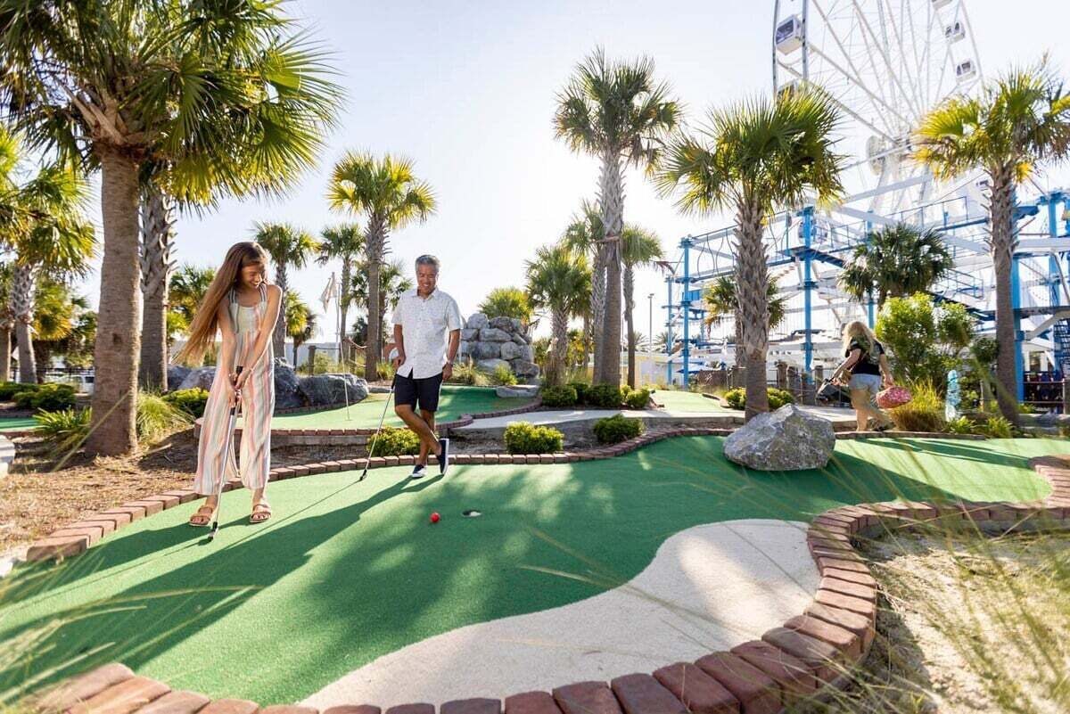 A miniature golf course with a green putting surface, palm trees, and a Ferris wheel in the background.