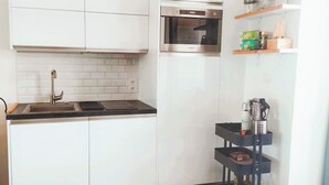 Cocina compacta con un lavamanos de acero inoxidable, microondas y horno empotrado. Hay armarios blancos y una encimera negra.