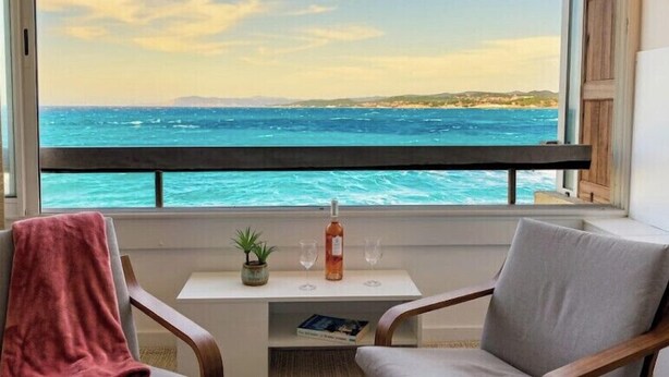 Un balcón con vista al mar, una botella, vasos, una planta y un libro sobre una mesa.