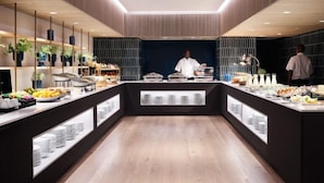 A modern breakfast buffet with a central serving area, surrounded by shelves with fruits, pastries, and beverages. - Hyatt Regency London Stratford (London)