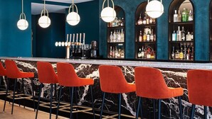 A bar area with red bar stools, a marble countertop, and a blue wall with shelves of liquor bottles. - Hyatt Regency London Stratford (London)