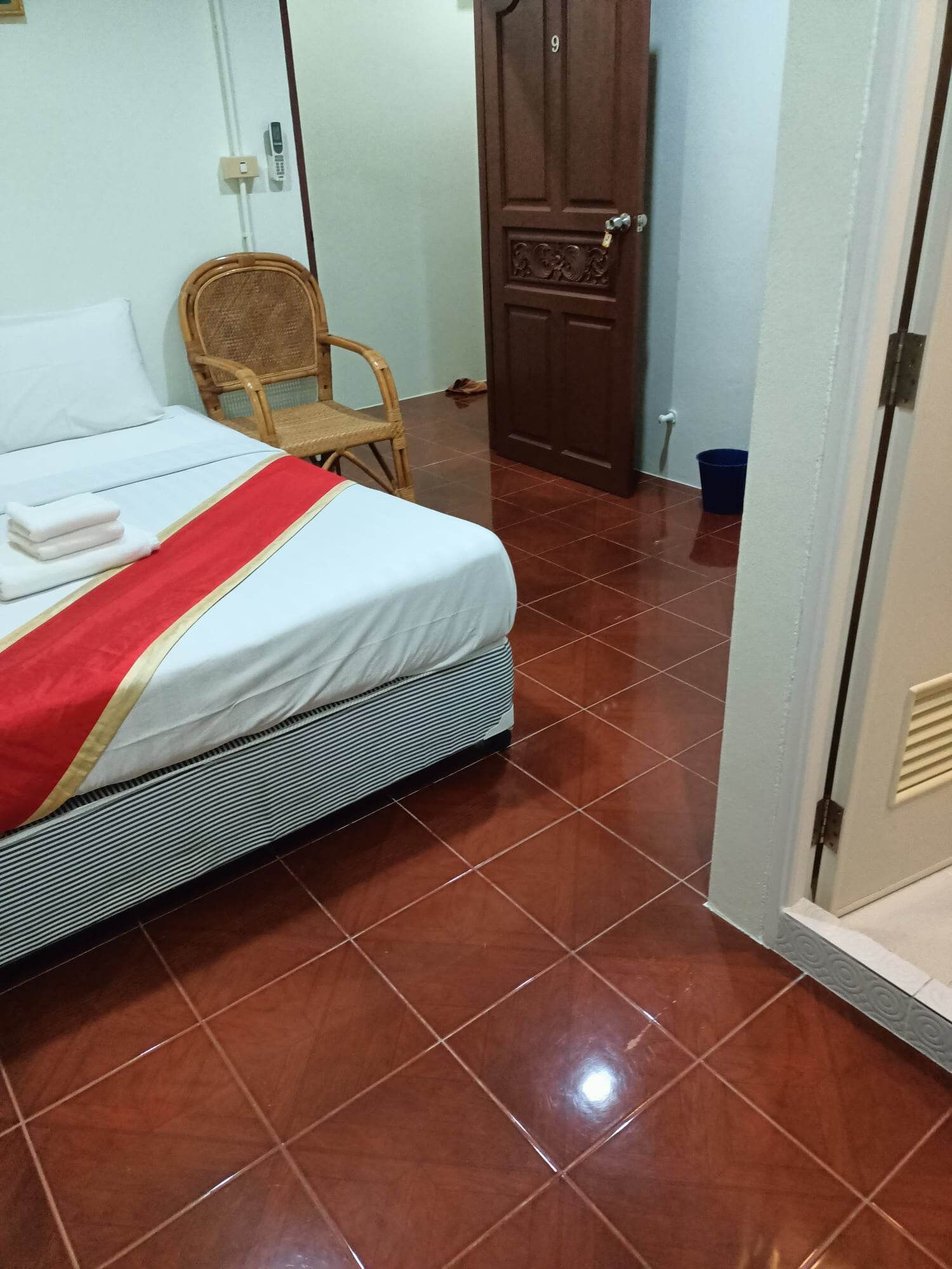 A neatly made bed with a red and gold bedspread, a wicker chair, and a wooden door.
