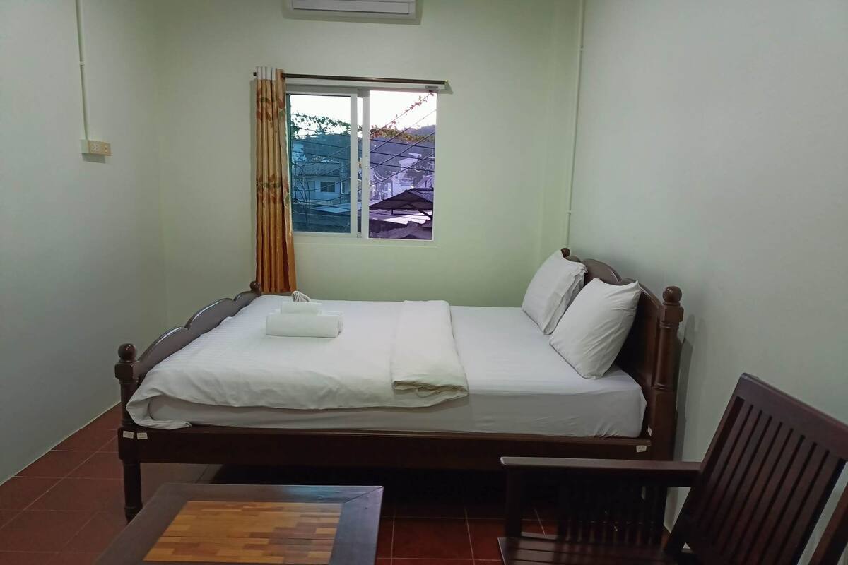Uma cama de solteiro com roupa de cama branca, cabeceira de madeira e janela com vista para edifícios.