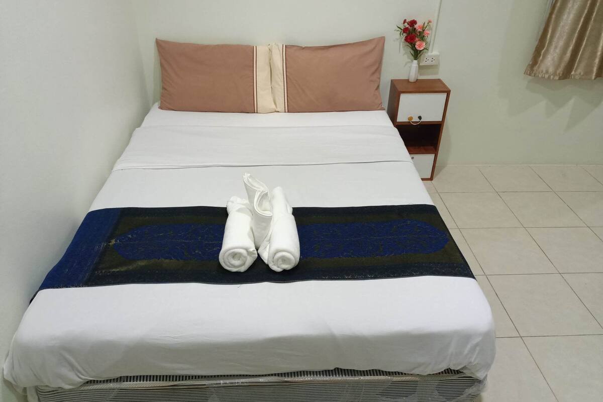 Uma cama bem arrumada com roupa de cama branca e bege, um cobertor listrado em azul e preto, e duas toalhas brancas enroladas.