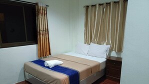 A single bed with a beige and blue bedspread, white pillows, and a rolled white towel on the footboard. There is a window with brown curtains, a brown headboard, and a wooden nightstand with a drawer. - Kata S.T House 2 (Karon)