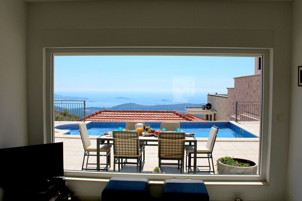 A room with a large window overlooking a swimming pool and a scenic view of the sea and hills.