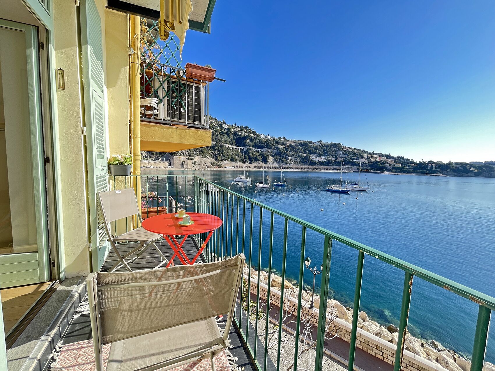 A balcony with a table and chairs, overlooking a calm sea with boats and a hilly landscape in the background.